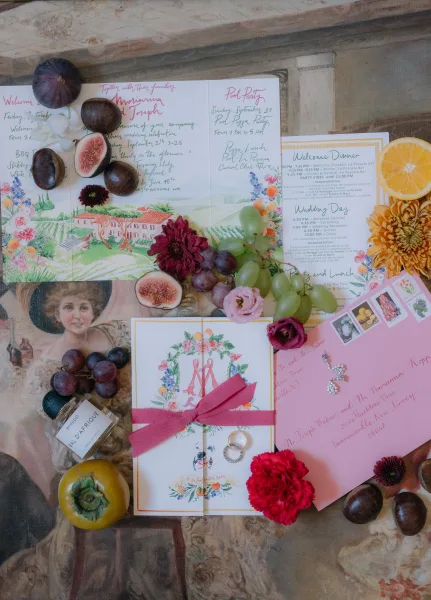 Invitation suite with watercolor wedding invitations, envelopes, ribbon, wax seal, stamps, rings, perfume, and fruit on vintage paper tabletop