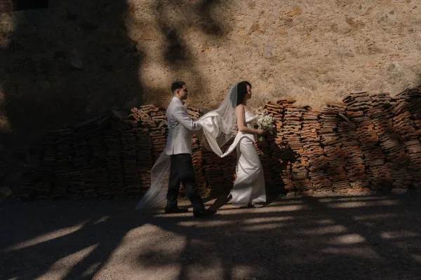 Couple portrait of bride and groom walking, groom holding veil as bride carries a white bouquet beside a stone wall in tree shadows