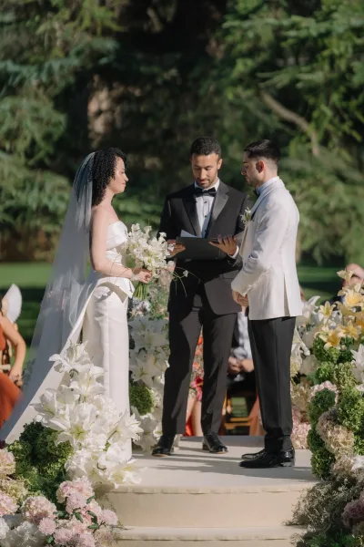 Wedding vows as bride in long veil holds lily bouquet beside groom in white tuxedo during outdoor wedding ceremony on garden lawn