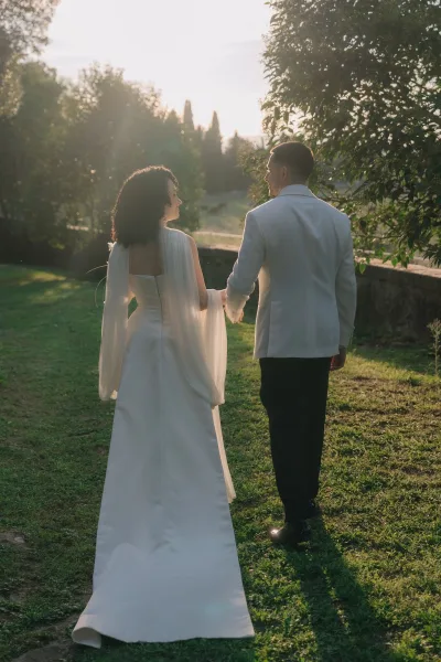 Couple portrait of bride and groom walking away holding hands, bride in a bridal cape and long train, by stone wall at sunset