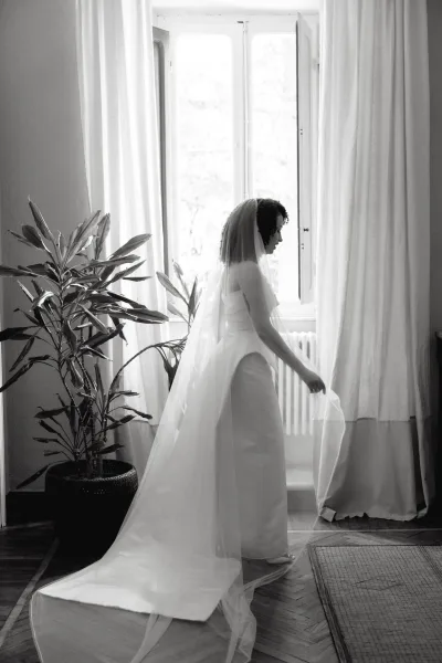 Bridal portrait of a bride by window in a strapless gown with long train, wearing a veil, softly backlit through sheer curtains indoors