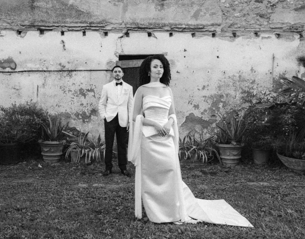 Couple portrait in a black and white wedding portrait, bride in draped-sleeve gown with train beside groom in tux by plaster wall doorway.