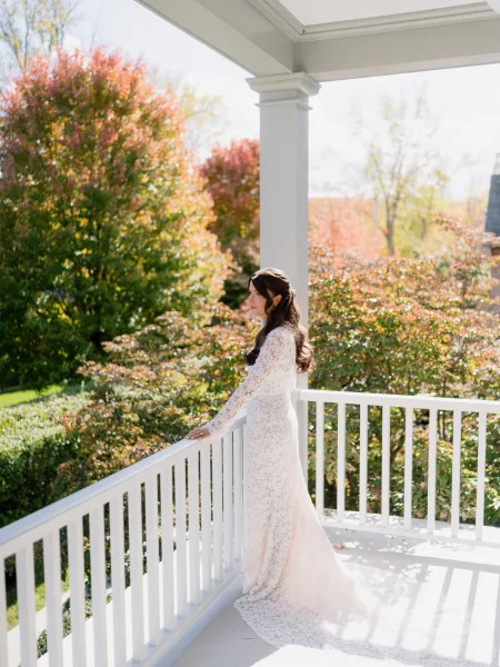 Bridal portrait of a bride in a long-sleeved gown on a white-railed porch, autumn garden trees behind her as she gazes away in soft natural light