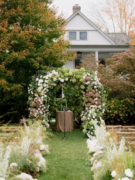 Ceremony arch anchors garden ceremony decor, adorned with hydrangeas on a lawn beneath autumn trees and near a house exterior.