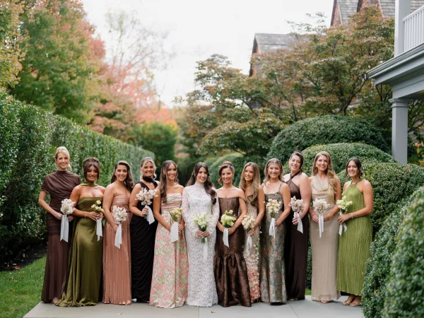 Bridesmaids group photo in mix and match bridesmaid dresses, holding ribbon-tied bouquets by garden hedges and fall trees.