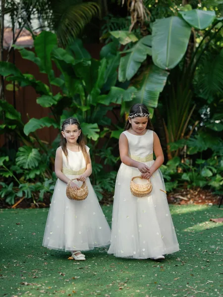 Flower girl moment during the wedding processional, with two girls carrying wicker baskets along a lawn framed by tropical plants.