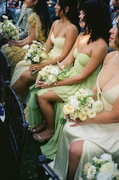Bridesmaid dresses in a mix and match bridesmaids lineup, bouquets in hand, seated by ceremony chairs on a grass lawn with guests.