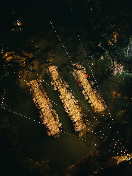 Outdoor reception setup with long banquet tables under string lights on a grass lawn bordered by garden trees, ready for an evening celebration.