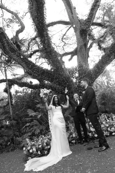 Ceremony moment at the wedding pronouncement as the bride and groom beam beneath a large tree canopy with string lights.