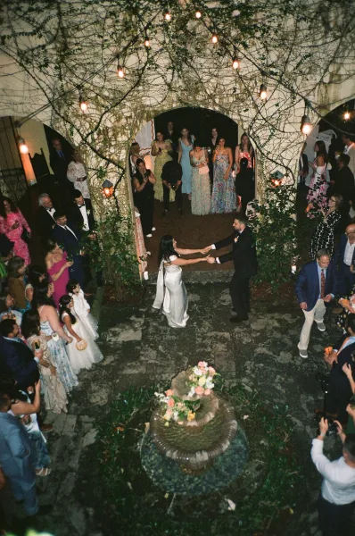 First dance, a couple dancing in a stone courtyard beneath warm string lights as guests surround the floor by a vine-covered wall.