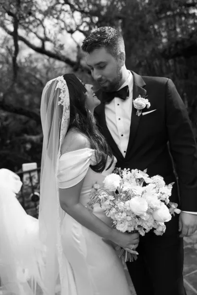 Couple portrait of a bride and groom embrace, her lace veil brushing his shoulder beneath garden trees along a stone path.