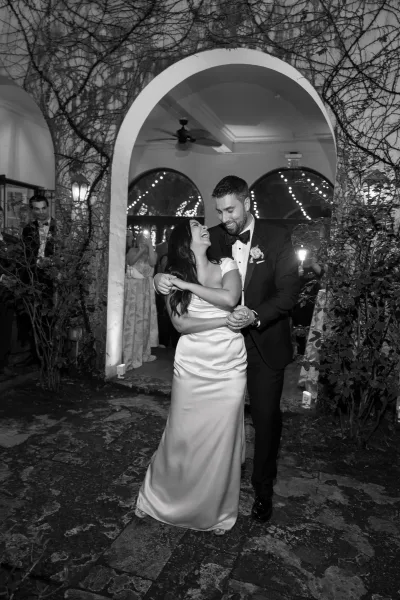 Wedding first dance with the bride and groom dancing beneath warm string lights on a stone patio by an arched doorway and vine-covered wall.