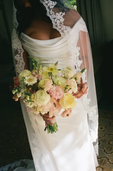 Bridal portrait of a bride holding a bouquet, lace veil draping softly in an indoor room with curtains and a patterned rug.