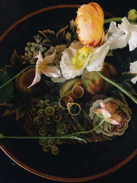 Wedding rings with engagement ring detail on a painted tray, framed by delicate florals over a dark tabletop for a moody feel.