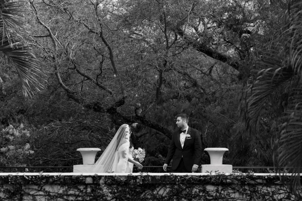 Wedding first look couple portrait as the bride walks toward the groom, lace veil trailing, against an ivy-covered garden terrace.