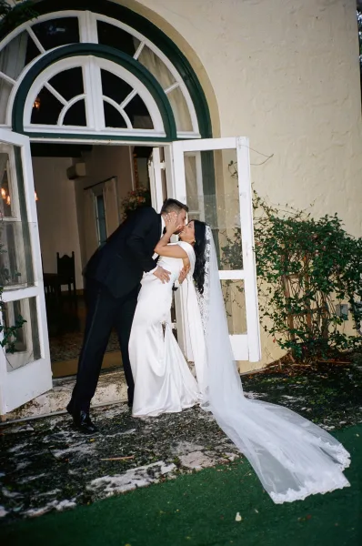 Wedding kiss portrait as a bride and groom kiss beneath an arched doorway, her long veil flowing by French doors and garden vines.
