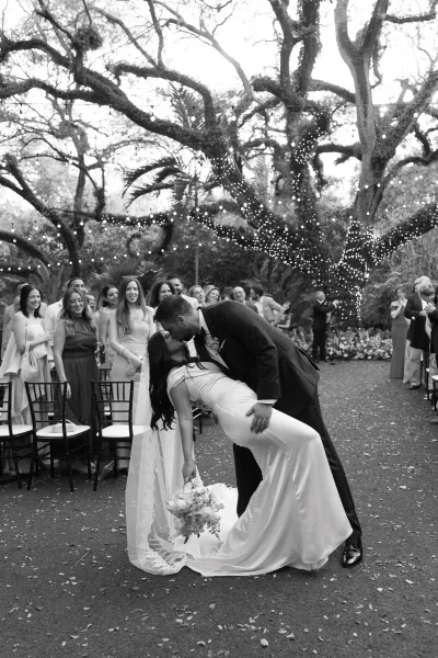 Wedding kiss portrait capturing a romantic dip kiss wedding photo on an outdoor garden aisle, string lights twinkling above the newlyweds.