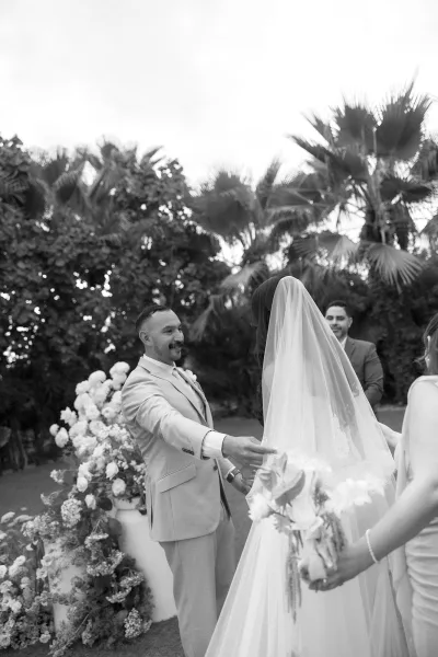 Ceremony moment as the groom greets the bride, her bridal veil trailing, amid palm trees and lush garden greenery.