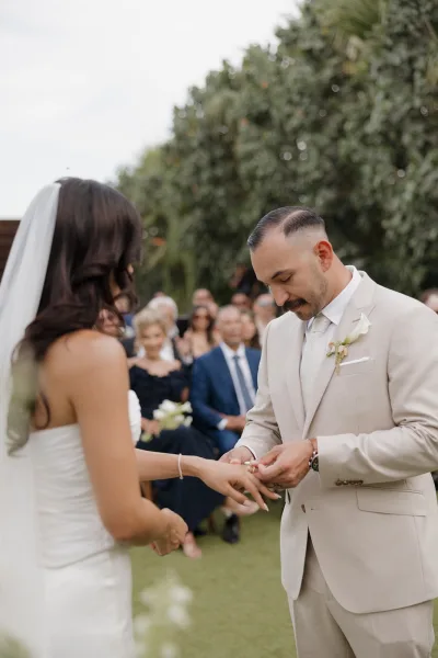 Ring exchange as the groom places the ring on the bride’s finger, her veil softly draped, amid garden palms and seated guests.
