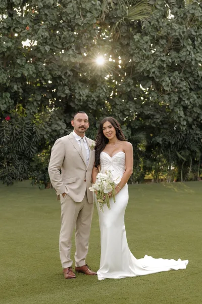 Couple portrait of a bride and groom standing as she holds a white bridal bouquet in a sunlit garden framed by palm leaves and green lawn