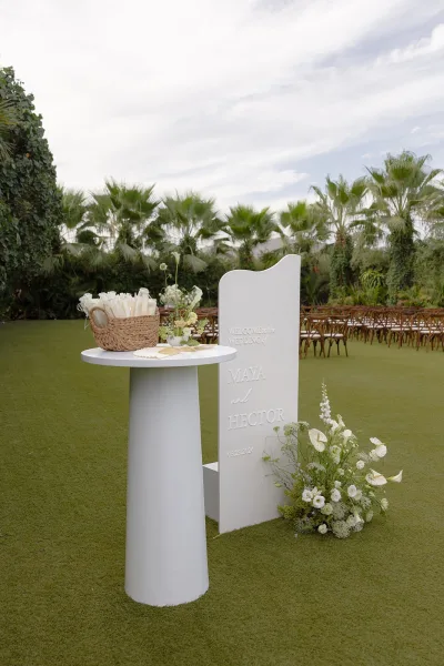 Wedding welcome sign serving as ceremony entrance decor beside a basket of fans on a lawn, with palm trees and ceremony chairs beyond.
