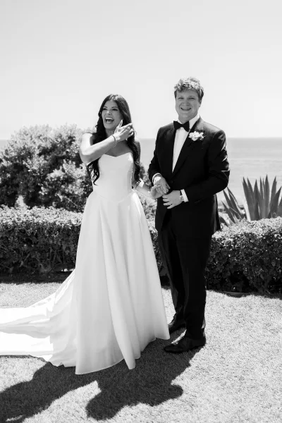 Couple portrait of bride and groom laughing and holding hands, strapless gown and tuxedo, with ocean horizon behind on lawn grass