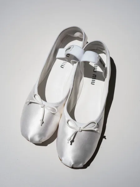 Bridal shoes in white bridal flats, satin with bow detail and elastic heel strap, arranged on a white surface with hard shadow