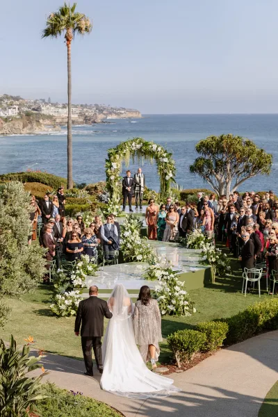 Wedding ceremony setup with a clear acrylic aisle as the bride in a long veil walks toward a white floral arch by the ocean cliffs
