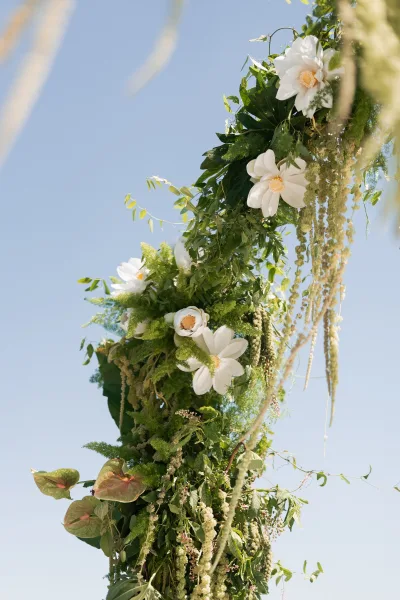 Wedding floral arch with white flowers, greenery, and hanging amaranthus trailing from tropical leaves against a bright blue sky backdrop