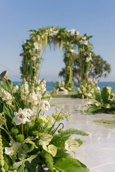 Ceremony arch decor with hanging greenery and white flowers, framing an oceanfront aisle runner under blue sky and distant trees