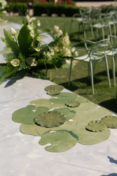 Reception tablescape with green wedding table decor, leaf-shaped placemats on white linen, white flowers and greenery centerpiece on a garden lawn