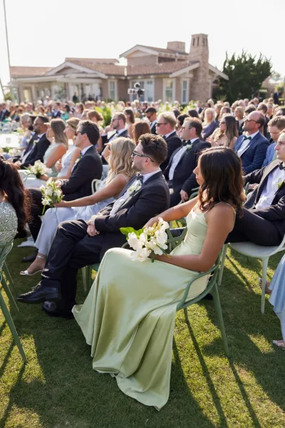 Wedding guests seated at ceremony in tuxedos and sage bridesmaid dresses, some with sunglasses, on green chairs on a sunny lawn
