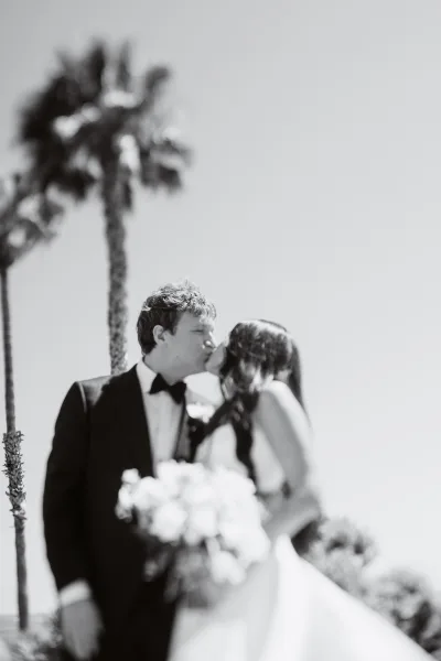Wedding kiss as bride and groom kiss in a strapless gown and tuxedo, holding a white rose bouquet with palm trees behind them