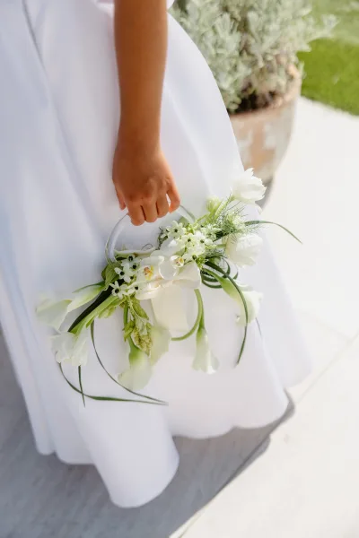 Bridal bouquet of white wedding bouquet blooms with lush greenery and trailing ribbon held against a bridal gown on an outdoor walkway