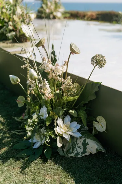 Ceremony florals with aisle meadow flowers in white blooms and lush greenery, accented by lotus pods and large leaves on an oceanfront lawn