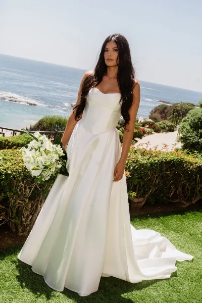Bridal portrait of a bride in a strapless satin wedding dress holding a white bouquet with greenery on a cliffside lawn overlooking the ocean