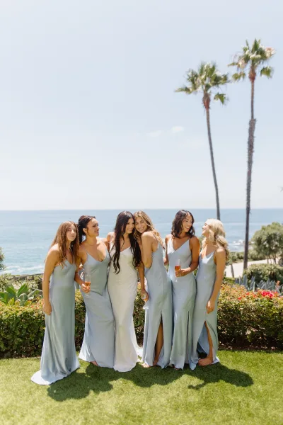 Bridesmaid group photo with bride and bridesmaids in matching bridesmaid dresses holding champagne flutes on an oceanfront lawn by palms