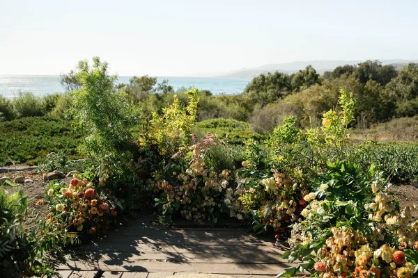 Ceremony altar decor with grounded wedding arch flowers in asymmetrical greenery and ground florals on a wood deck with ocean view