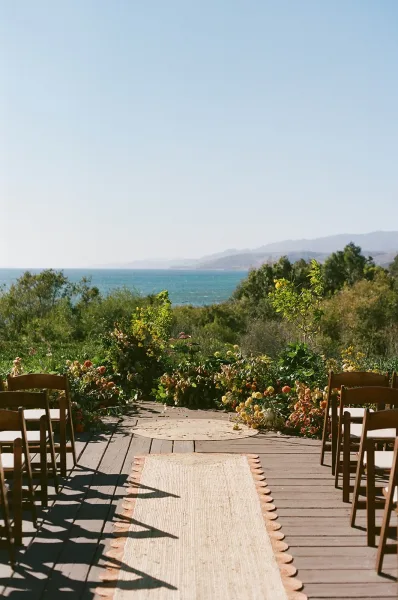 Ceremony setup for an outdoor wedding ceremony with wood folding chairs, aisle runner, and floral arrangements on a deck with ocean view