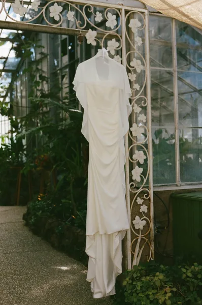Wedding dress hanging on a hanger, draped softly against a wrought iron gate with greenhouse windows and garden plants behind