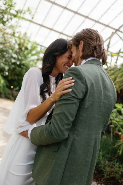 Couple portrait of bride and groom embracing with a forehead touch, her gold jewelry and long-sleeve dress under a greenhouse ceiling