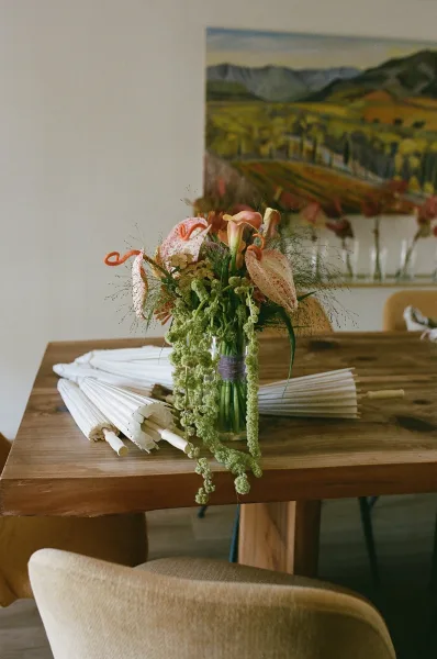 Wedding bouquet with calla lily bouquet blooms and hanging amaranthus in a glass vase on a wood table, with white folding fans nearby