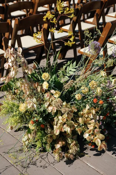 Ceremony aisle flowers in a meadow-style ground arrangement with yellow roses and wildflowers beside wooden folding chairs on a deck