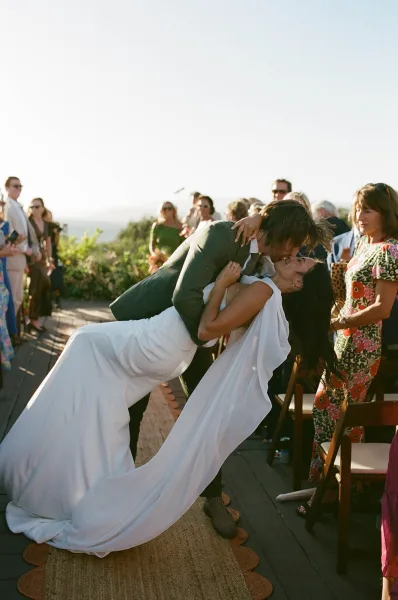 Wedding kiss portrait of bride in a draped cape and groom in suit dipping mid-aisle on an outdoor terrace, guests seated beside runner