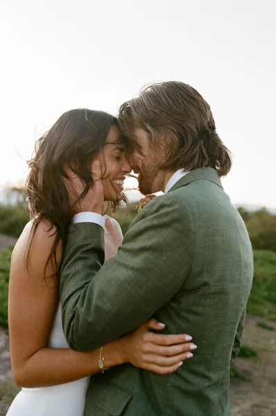 Couple portrait of a wedding couple embrace with foreheads touching, groom in a green suit holding bride’s face against open greenery and sky