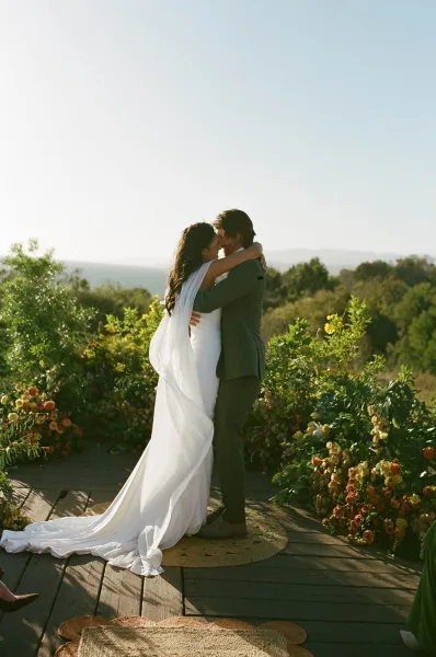 Wedding kiss portrait of bride and groom kiss on a wooden deck, her long veil blowing with hills and trees behind them in soft light