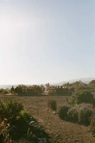 Outdoor wedding ceremony with an aisle runner between wood chairs and floral arrangements, overlooking the ocean view from a hillside setting