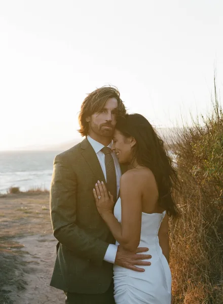 Couple portrait of bride and groom embrace on a coastal cliff by the ocean, bride in a strapless wedding dress with drop earrings in tall grass