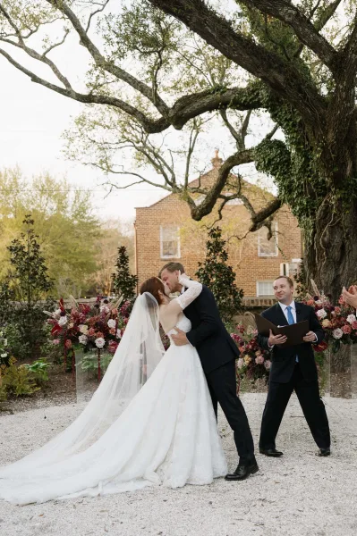 Ceremony kiss as the bride and groom kiss beneath a large tree, flanked by clear acrylic flower stands in a lush garden setting.