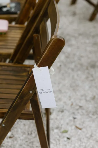 Reserved seating sign used as a ceremony chair sign, embossed florals on a paper tag, tied to a wooden chair over a gravel aisle.
